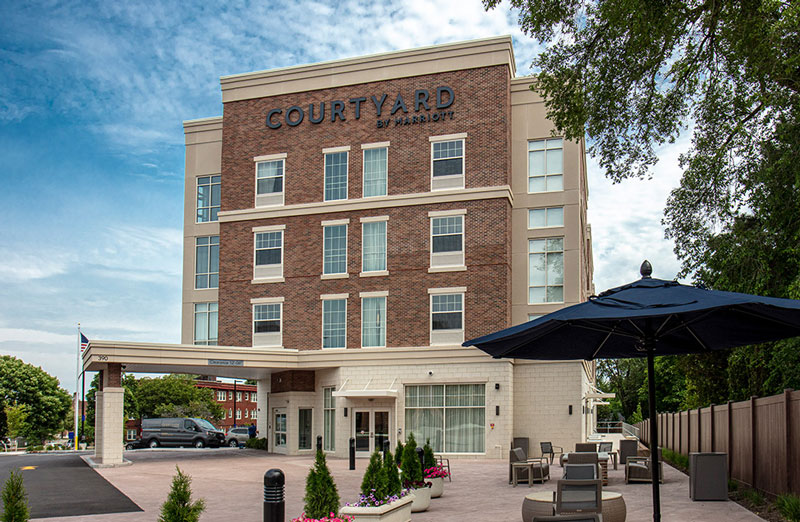 Exterior view of a Courtyard by Marriott hotel building with modern architecture, outdoor seating area, large umbrella, and surrounding greenery under a partly cloudy sky.