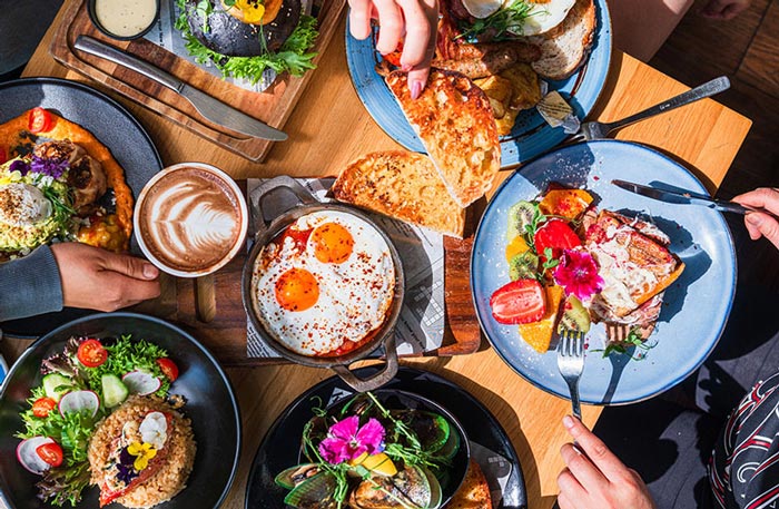 A vibrant brunch spread on a wooden table featuring sunny-side-up eggs, fresh salads, toast, tropical fruits, a latte with latte art, and various gourmet dishes being shared by multiple hands.