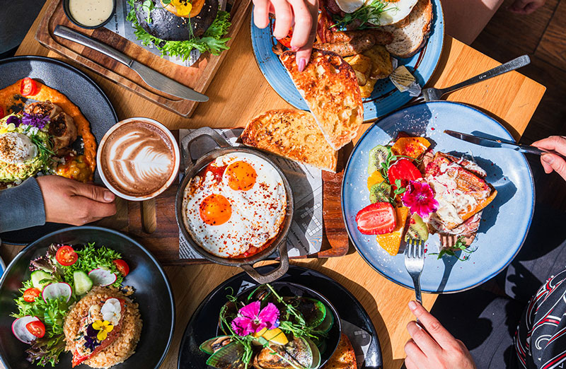 A vibrant brunch spread on a wooden table featuring sunny-side-up eggs, fresh salads, toast, tropical fruits, a latte with latte art, and various gourmet dishes being shared by multiple hands.