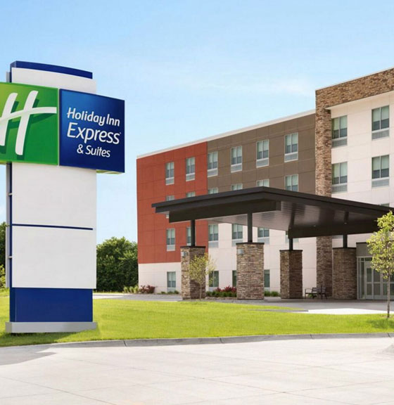 Modern exterior of a Holiday Inn Express & Suites hotel in Mechanicsburg, featuring a colorful facade with red, brown, and white panels, a large branded sign, and a covered entrance set against a clear blue sky.
