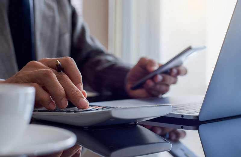 Close-up of a businessperson using a calculator while holding a smartphone, with a laptop and coffee cup on a reflective desk, suggesting multitasking and financial work.