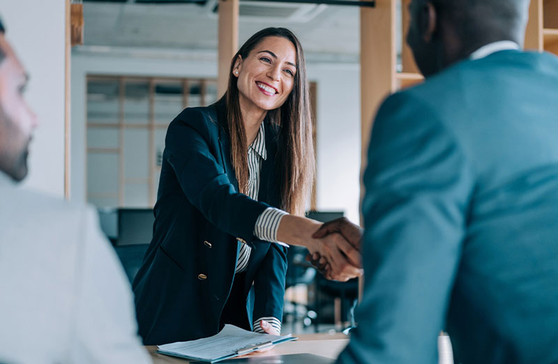 Professional woman smiling and shaking hands with a colleague in a modern office setting, symbolizing a successful business meeting or job interview.