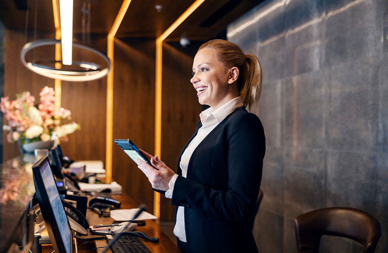 Smiling hotel receptionist or front desk professional in a modern, stylish lobby, holding a tablet while assisting guests using multiple computer monitors.
