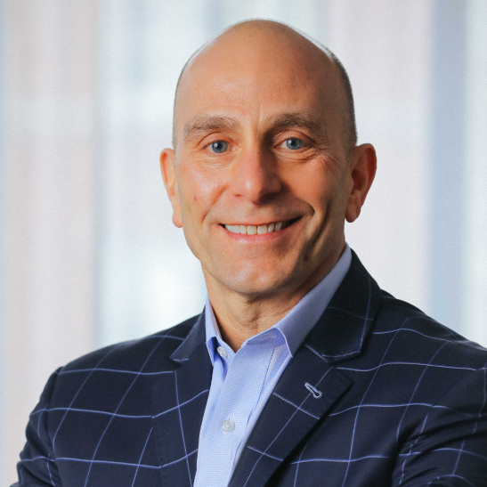 John Kattato - Confident, smiling business professional in a windowpane-patterned blazer and light blue shirt, posing in front of softly blurred curtains.