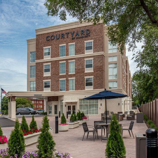 Front view of a Courtyard by Marriott hotel with a landscaped outdoor seating area, patio tables, large umbrella, and lush greenery under a partly cloudy sky.