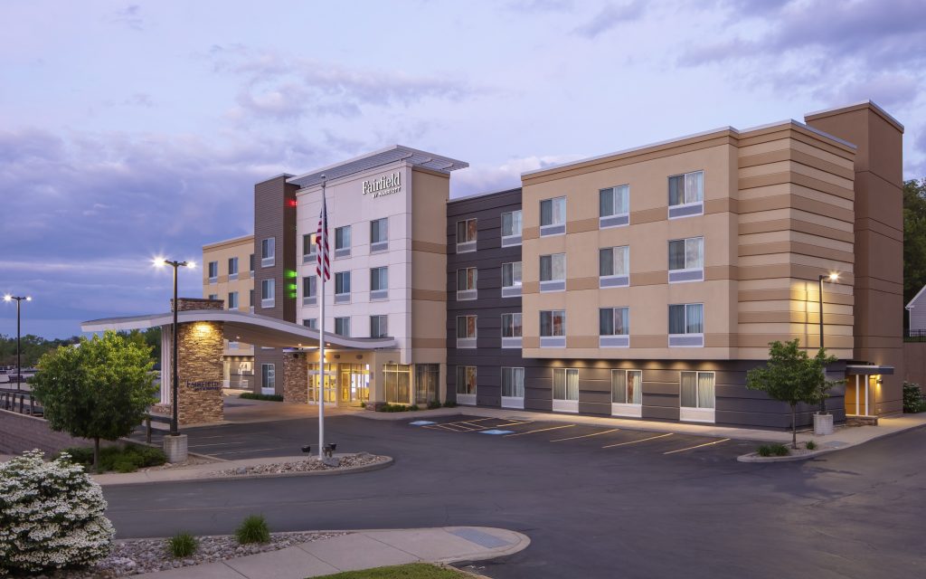 Exterior view of Fairfield Inn & Suites by Marriott featuring a modern four-story building, an American flag, and a parking lot at dusk with scattered clouds.