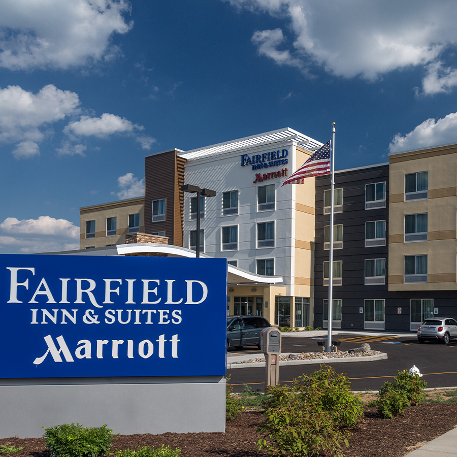 Front view of Fairfield Inn & Suites by Marriott in Geneva, featuring a modern four-story building, large blue brand signage, American flag, and neatly landscaped grounds under a partly cloudy sky.