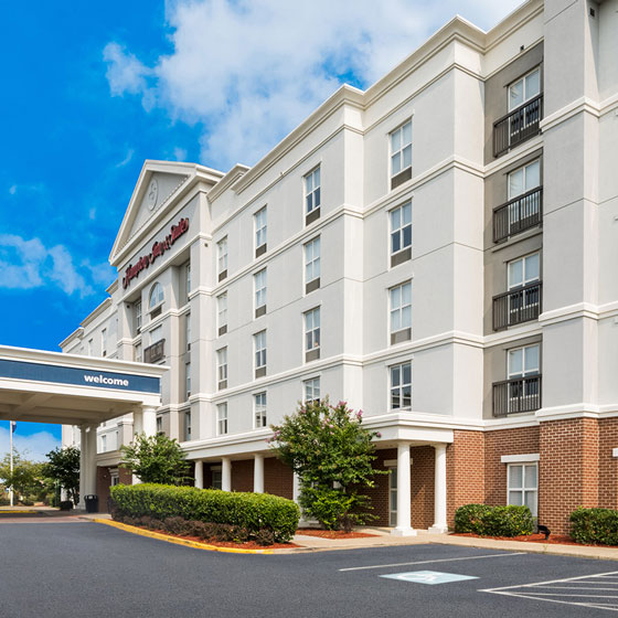 Exterior of Hampton Inn & Suites in Fredericksburg, featuring a five-story building with a brick and stucco facade, landscaped entrance, and a covered driveway under a bright blue sky.
