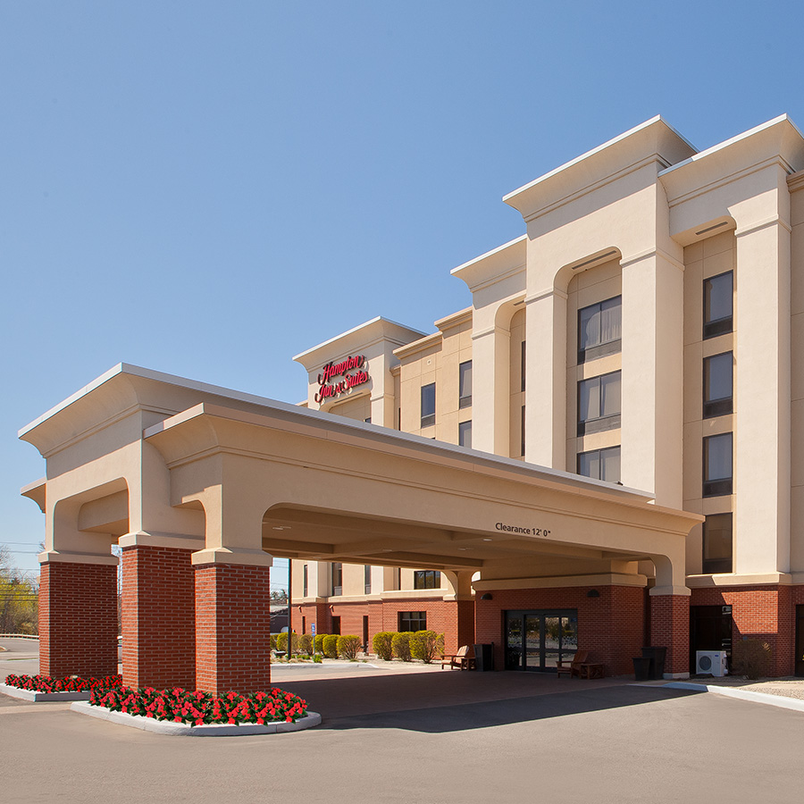 Front exterior of Hampton Inn & Suites in Plattsburgh, NY, showcasing a large covered entrance, beige and brick facade, manicured landscaping, and clear blue sky.