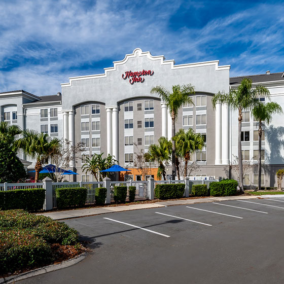 Front exterior of a Hampton Inn hotel with grand white columns, palm trees, and a gated outdoor area with blue umbrellas, set against a blue sky with light clouds.