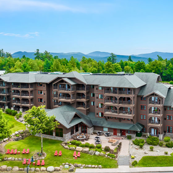 Aerial view of a rustic-style resort building surrounded by lush greenery and mountains, featuring balconies, outdoor seating areas, red Adirondack chairs, and landscaped grounds.