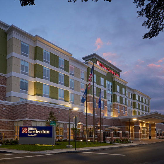 Exterior of Hilton Garden Inn in Corning, NY, at dusk, with illuminated signage, American and state flags, and a well-lit entrance against a colorful evening sky.