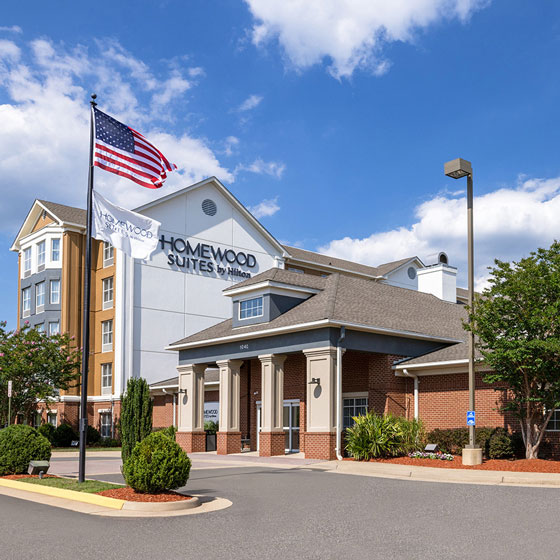 Exterior of Homewood Suites by Hilton in Fredericksburg, VA, featuring a traditional-style entrance, brick and siding facade, American and brand flags, and neatly landscaped surroundings under a bright blue sky.