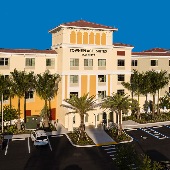 Exterior of TownePlace Suites by Marriott in Estero, featuring a Mediterranean-inspired design with warm-toned facade, palm trees, and a well-maintained parking area under a clear blue sky.